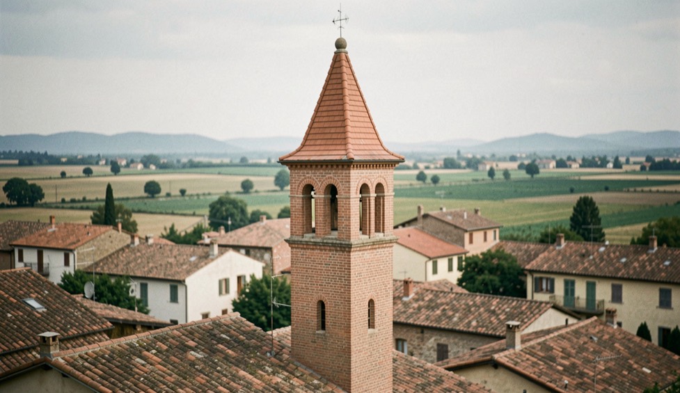 Immagine illustrativa: campanile in laterizio isolato tra i tetti di un paese di pianura, cella campanaria a bifore e cuspide in cotto